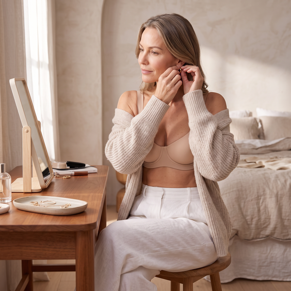 Woman putting on a gold earring at her vanity in the Crème nude beige Kischt Camille balconette bra with a cardigan over her shoulders