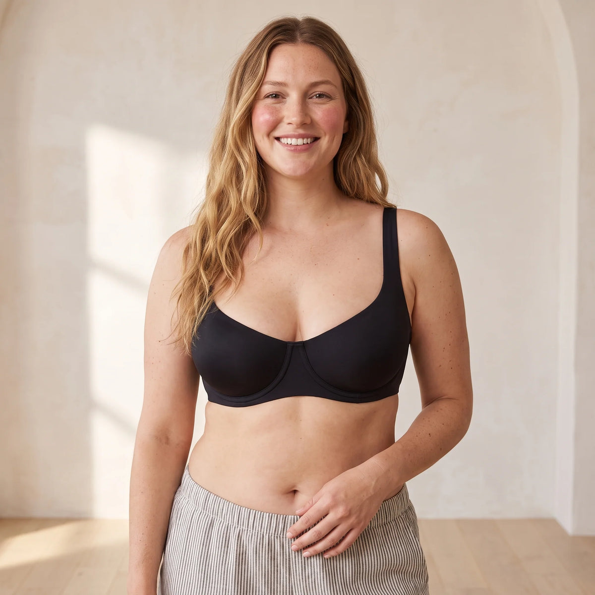 Smiling mum with wavy blonde hair wearing the Kischt Camille unlined balconette underwire bra in Noir black with striped linen trousers