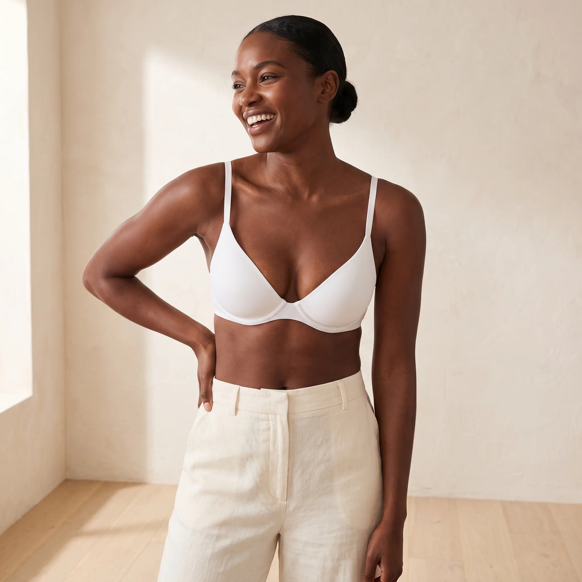 Laughing woman with a low bun wearing the Kischt Juliette unlined plunge underwire bra in Blanc white with cream linen trousers